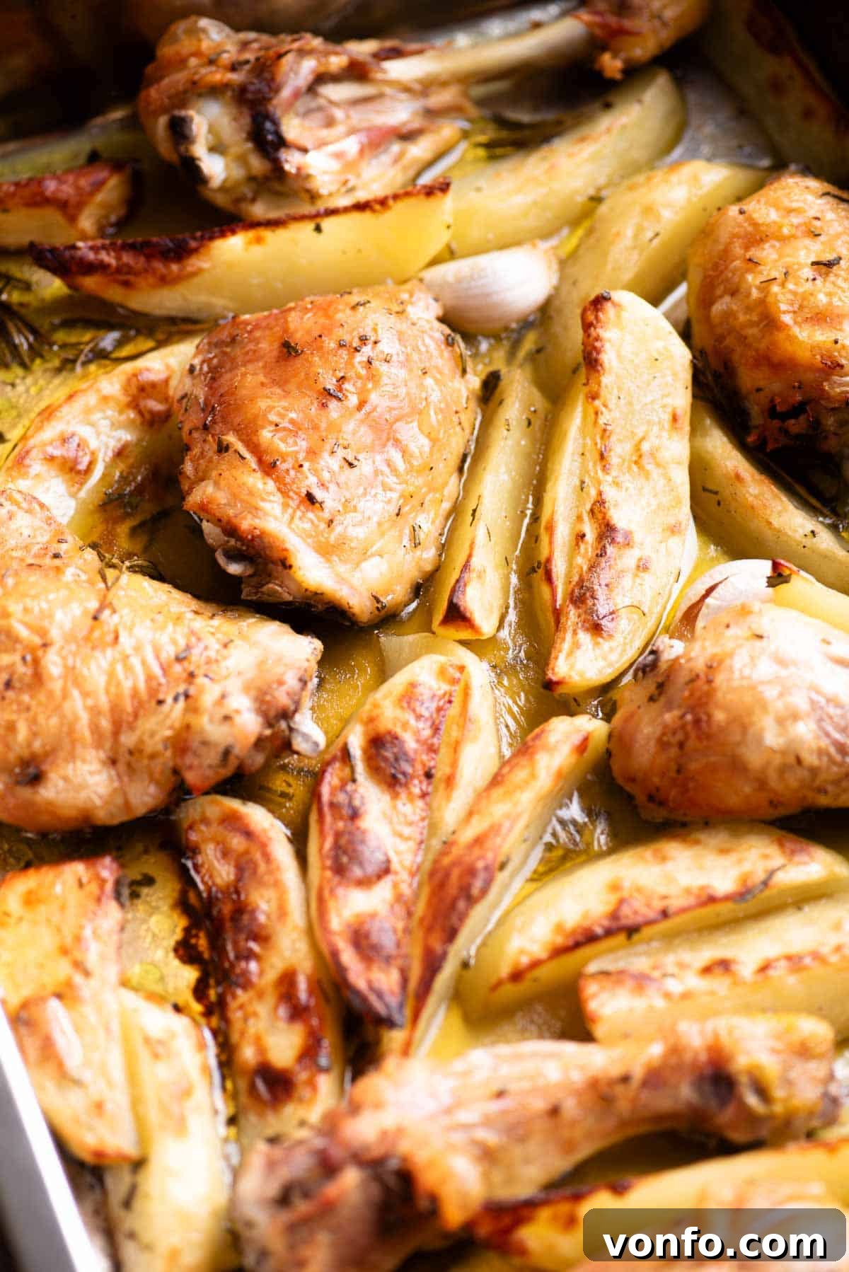 A close up side shot of roasted chicken thighs and drumsticks, glistening with juices, alongside tender potato wedges and fragrant rosemary in a dark roasting tray.