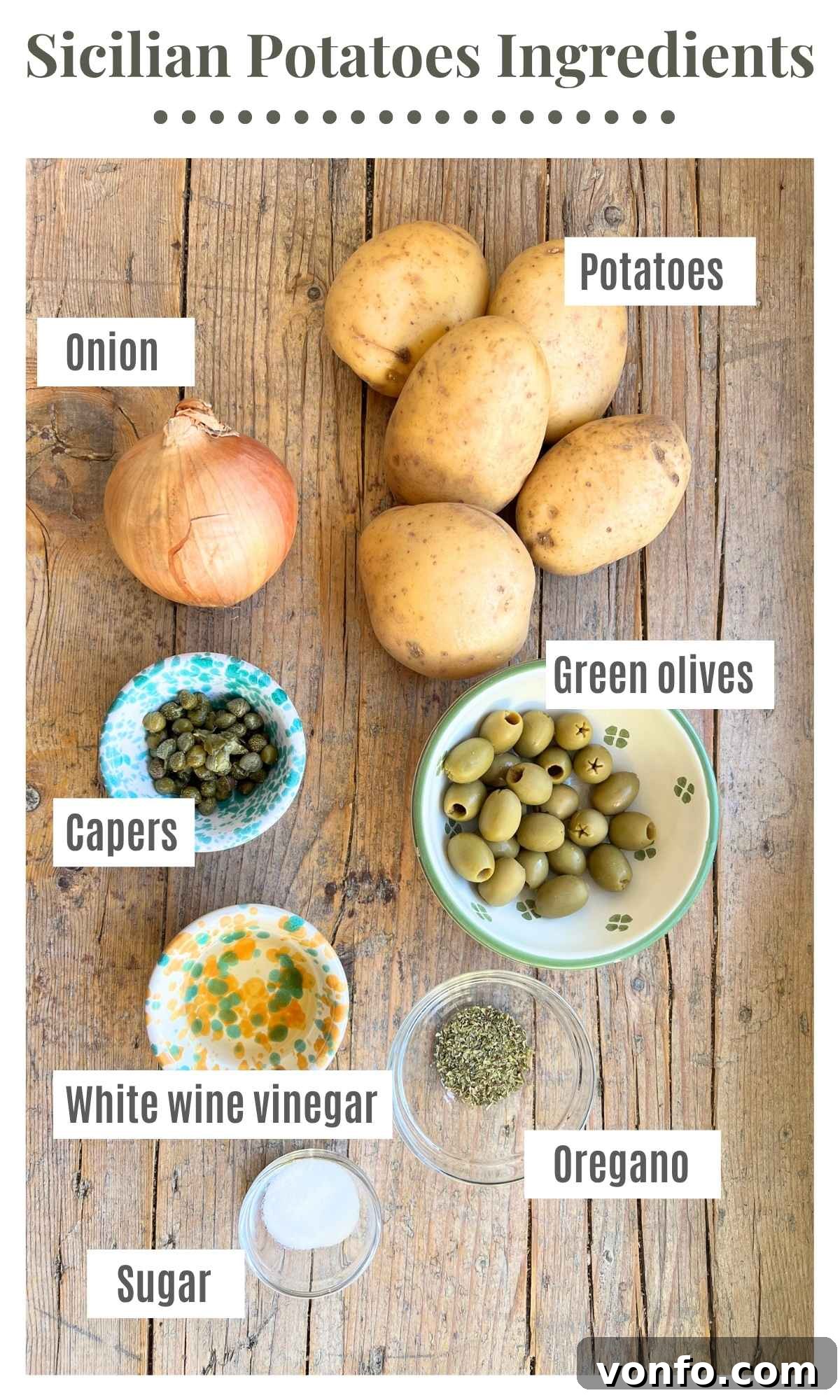 An overhead shot of all the fresh ingredients needed to make a traditional Sicilian potato side dish. Laid out are potatoes, onions, green olives, capers, white wine vinegar, sugar, and dried oregano.