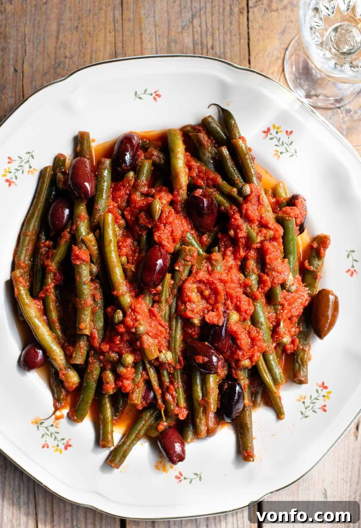 Zesty Green Beans with Olives and Capers 2 A close up of vibrant green beans in a rich red tomato sauce with briny olives and capers, served on a beautiful white floral plate against a rustic wooden background, ready to be enjoyed.