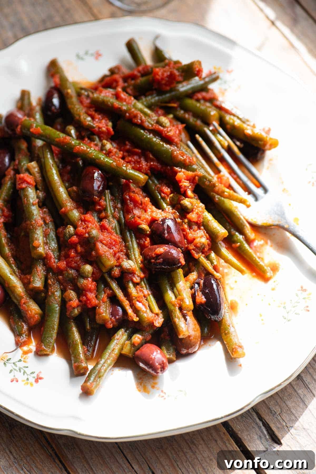 Zesty Green Beans with Olives and Capers 4 A side shot of perfectly cooked green beans with a rich tomato sauce, briny olives, and tangy capers, beautifully arranged on a white plate with a fork resting invitingly at the side.