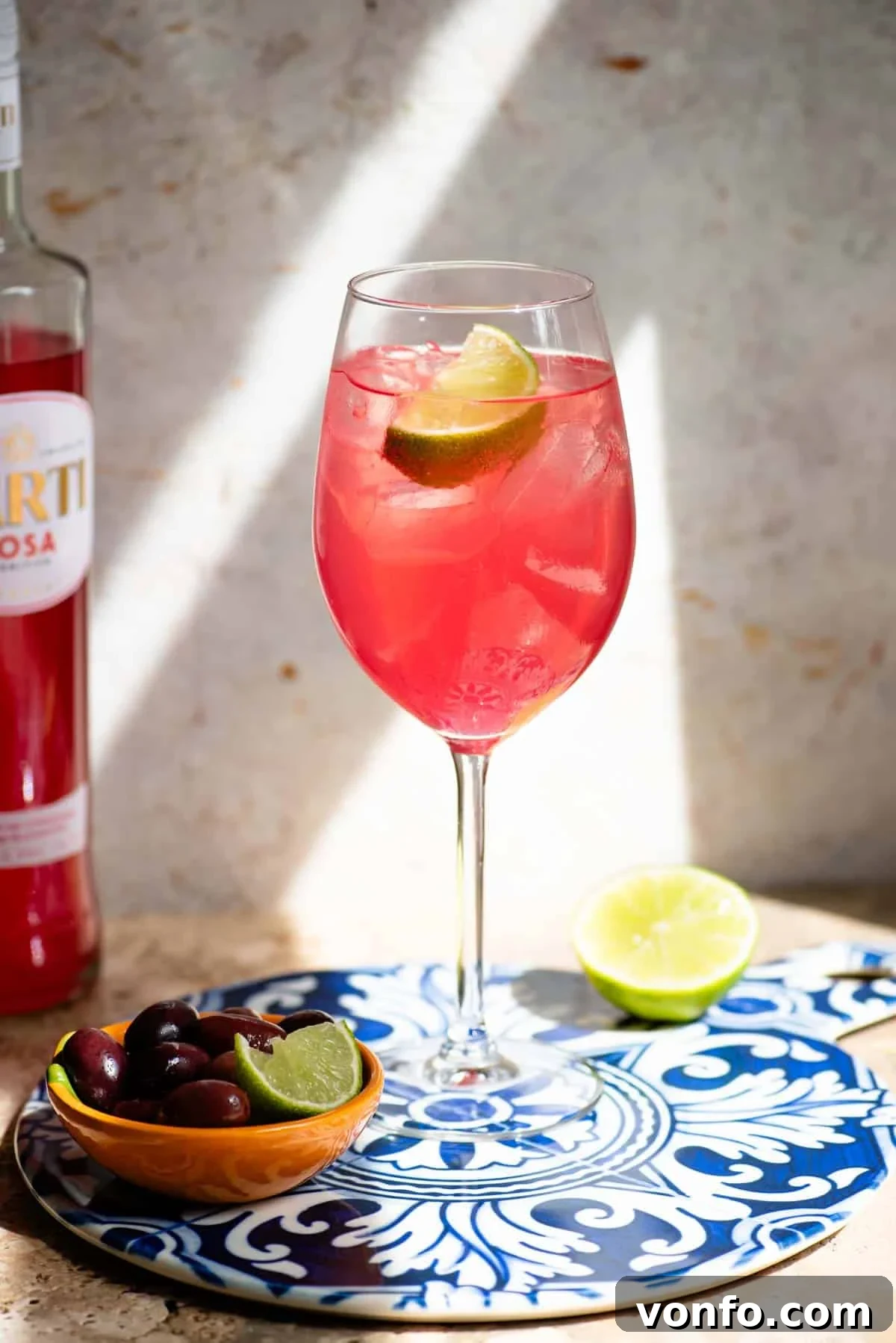 Sarti Spritz Fruity Italian Aperitif Cocktail 38 A Sarti Spritz in a wine glass with ice and a slice of lime sitting on a blue ceramic serving board with olive at the side.