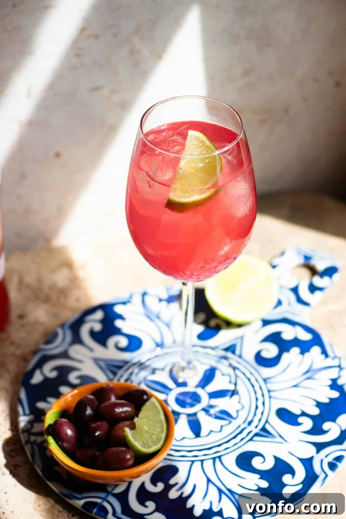 A close up of a sarti spritz with a wedge of lime on a blue patterned ceramic board with olives at the side.