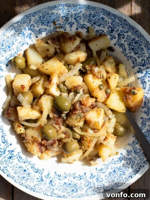 An overhead shot in direct sunlight of Sicilian potatoes in a blue bowl with olives and capers.