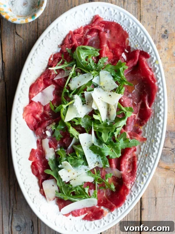 An overhead shot of beef carpaccio on a white oval serving plate topped with arugula and Grana Padano cheese.