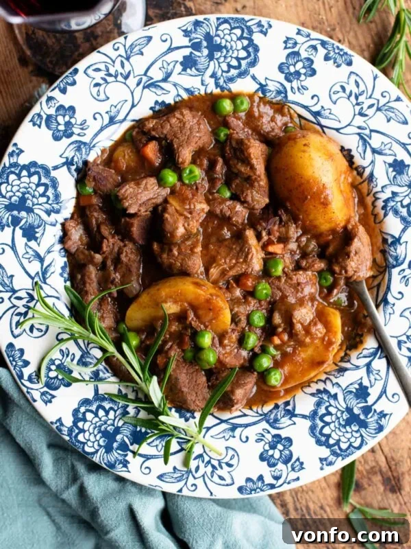 Italian beef stew with potatoes and peas in a blue and white floral bowl with rosemary and red wine at the side.