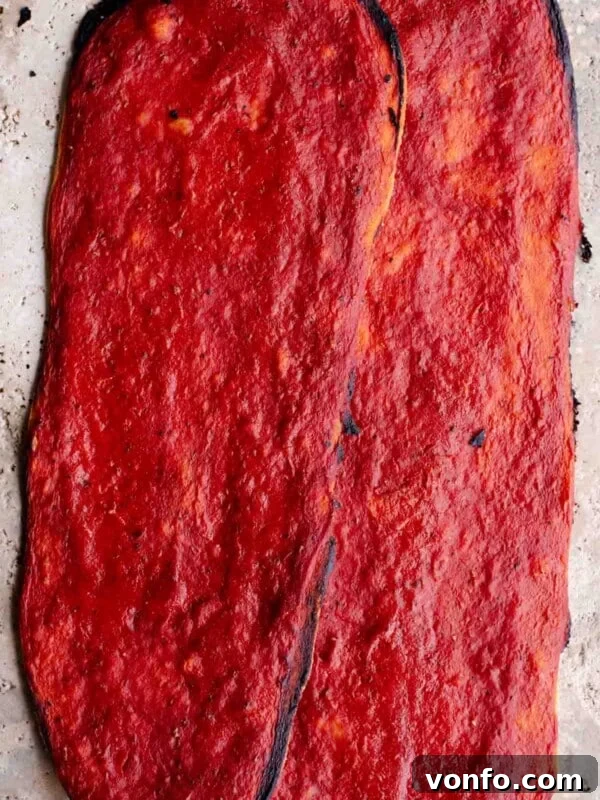 Mediterranean Pizza with Olives, Anchovies & Capers 7 Two long thin and crispy pizzas covered in tomato sauce sitting on a travertine surface.