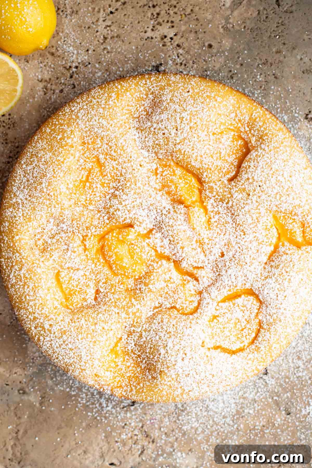 An overhead shot of a rustic Italian cake on a travertine surface with lemons at the side. The cake has dollops of pastry cream and powdered sugar.