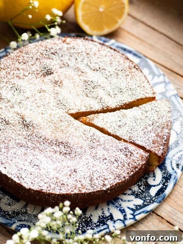 A white torta caprese on a blue ceramic board with fresh lemons behind it and small white flowers around.