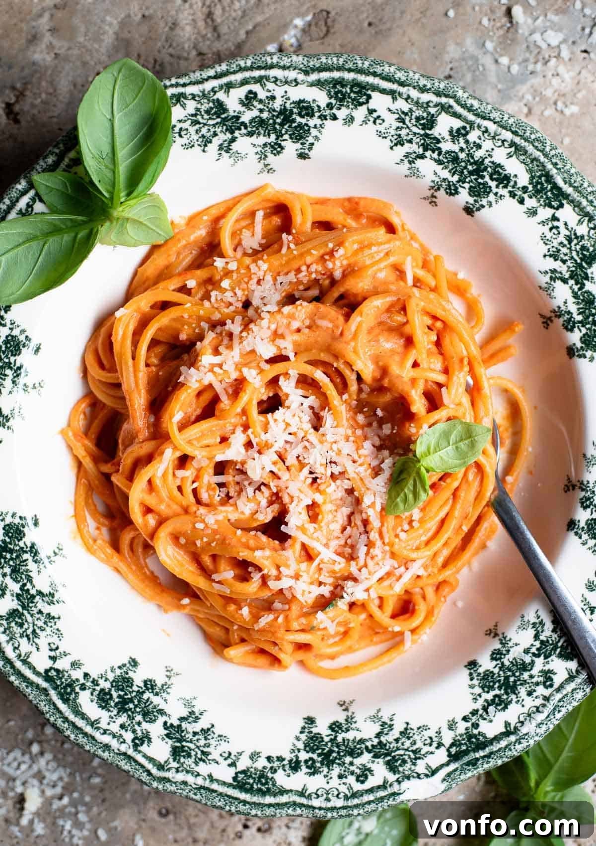 An overhead shot of roasted cherry tomato and ricotta spaghetti in a green floral bowl with basil scattered around.
