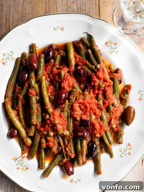 A close up of green beans in tomato sauce with olives and capers on a white floral plate and rustic wooden background.